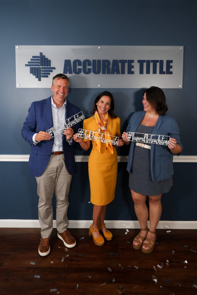 Three Accurate Title team members smiling and holding AskForAccurate and PoweredByTouchstone signs in the Accurate Title office